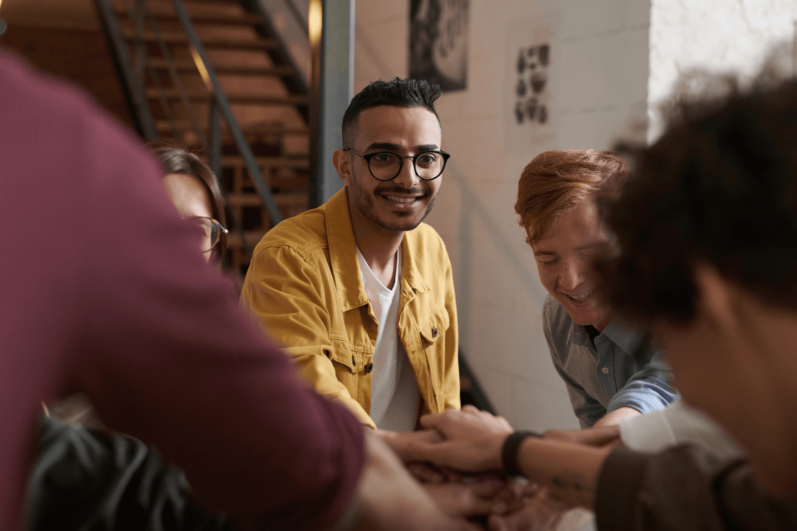 A group of students stands around a table, cheerfully placing their hands together in a gesture of teamwork and camaraderie, with a background featuring a staircase and wall decor.