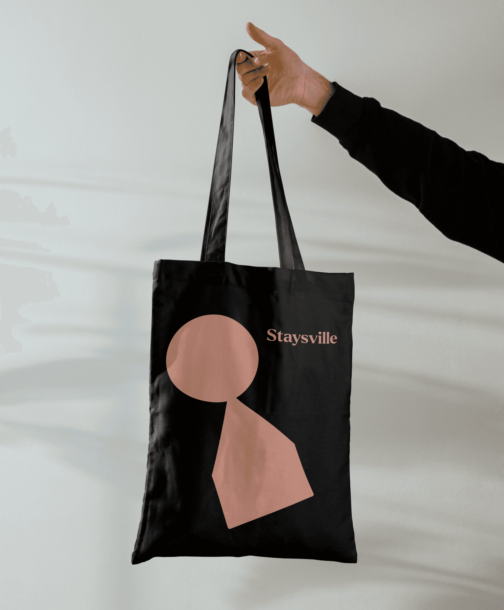 A hand holding a black totebag with the Staysville logo on it in pink on a white background.. 