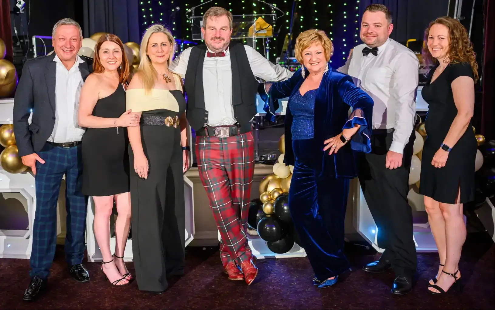 A group of seven people dressed in formal attire pose happily at an event, surrounded by black and gold balloons and festive decorations.