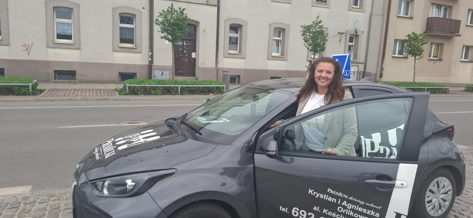 Agnieszka Zawolik – Instruktorka uśmiechnięta przy otwartych drzwiach szarego samochodu nauki jazdy.