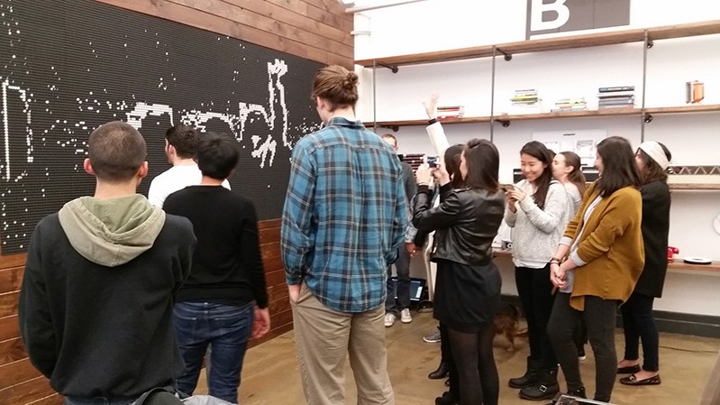 A photo of a group of students standing in an exhibition room, while looking at a blackboard with some white drawing.