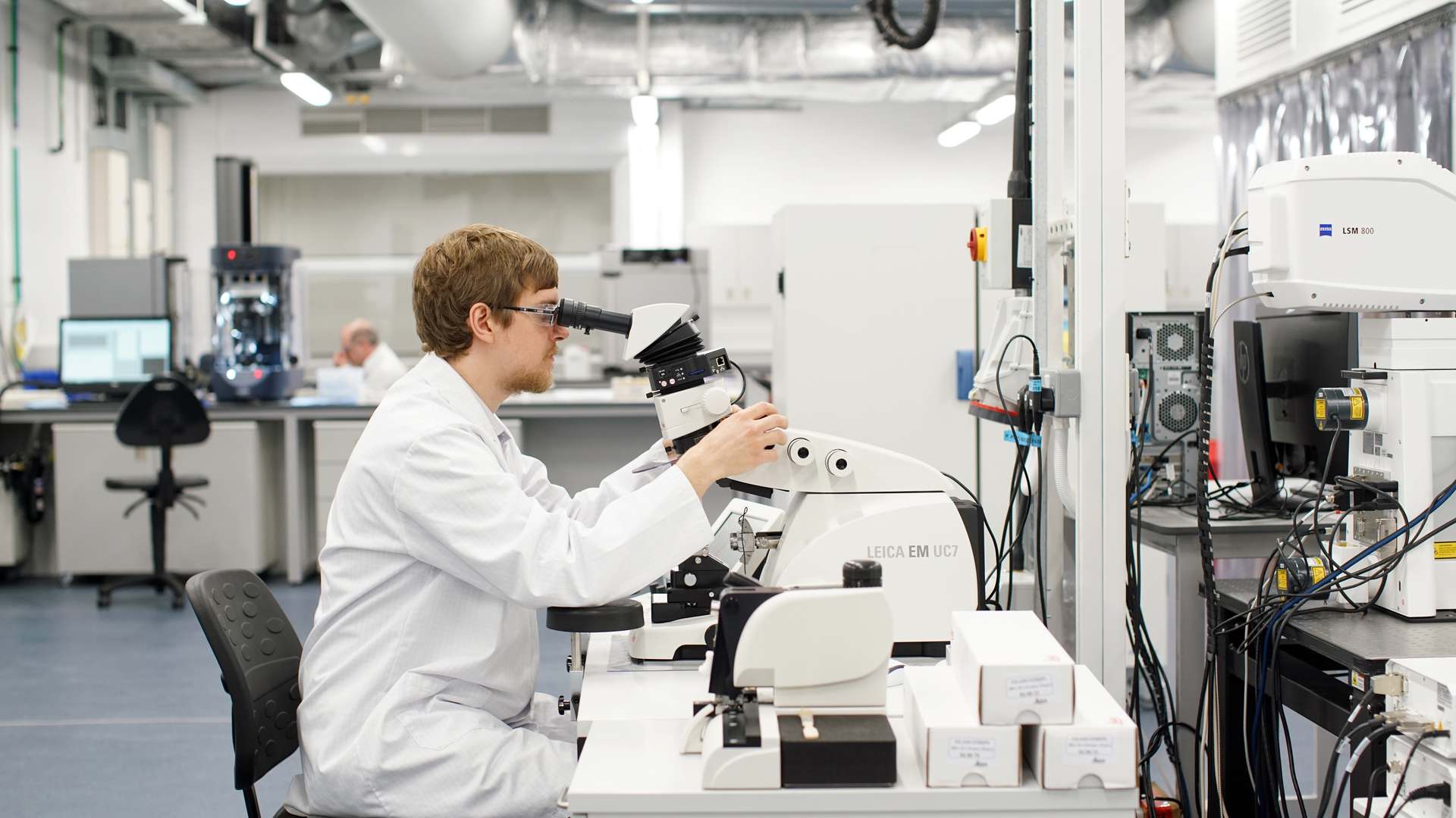 Male scientist in a white lab coat looking into a microscope at a bench in a large bright laboratory