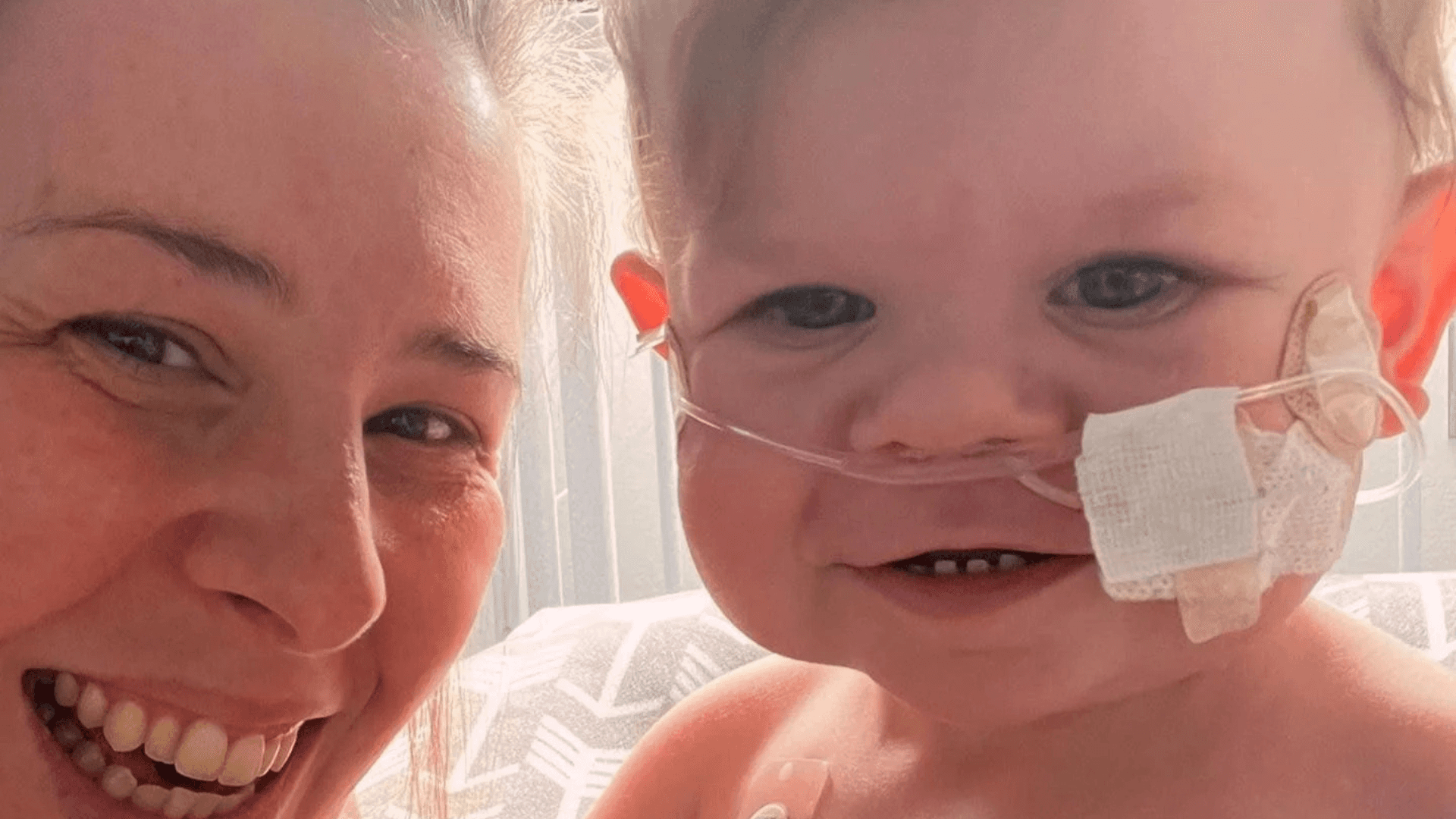Smiling mother and baby taking a close-up photo together, with the baby wearing a nasal cannula and medical tape on the cheek