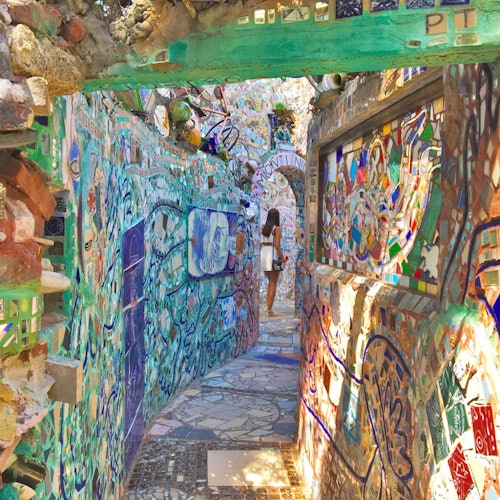 A narrow alleyway adorned with vibrant mosaics and sculptures, with a person walking through it, carrying a white bag.