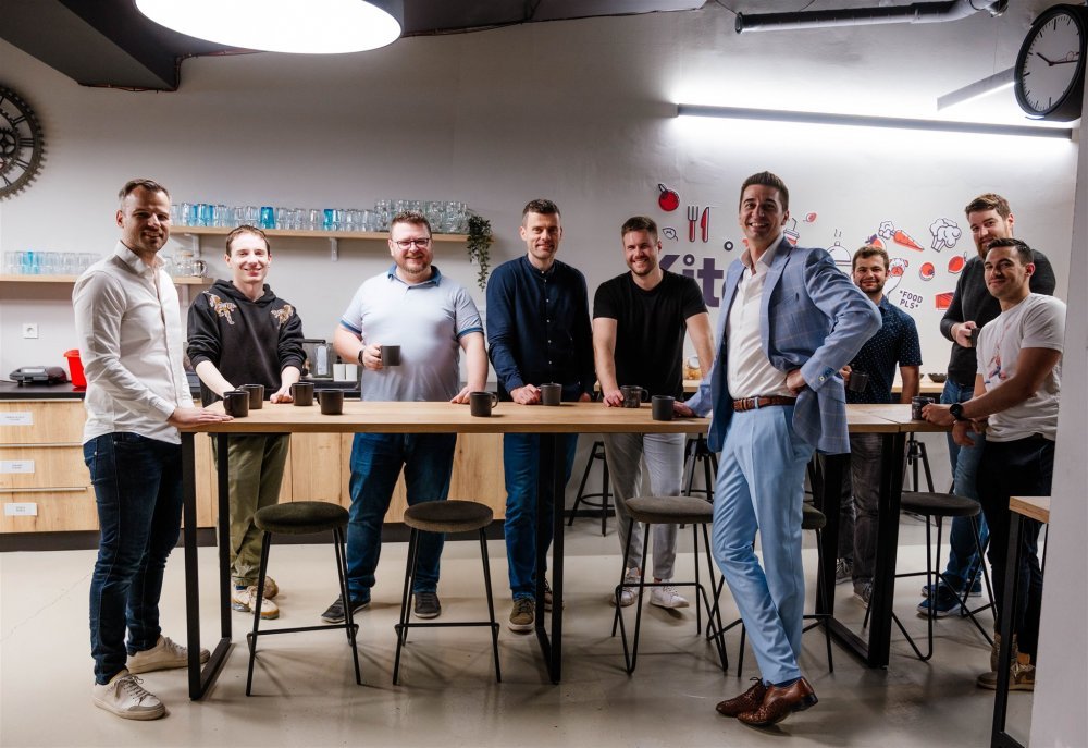 Man and woman standing together at a desk with a laptop and notebooks, smiling at the camera.