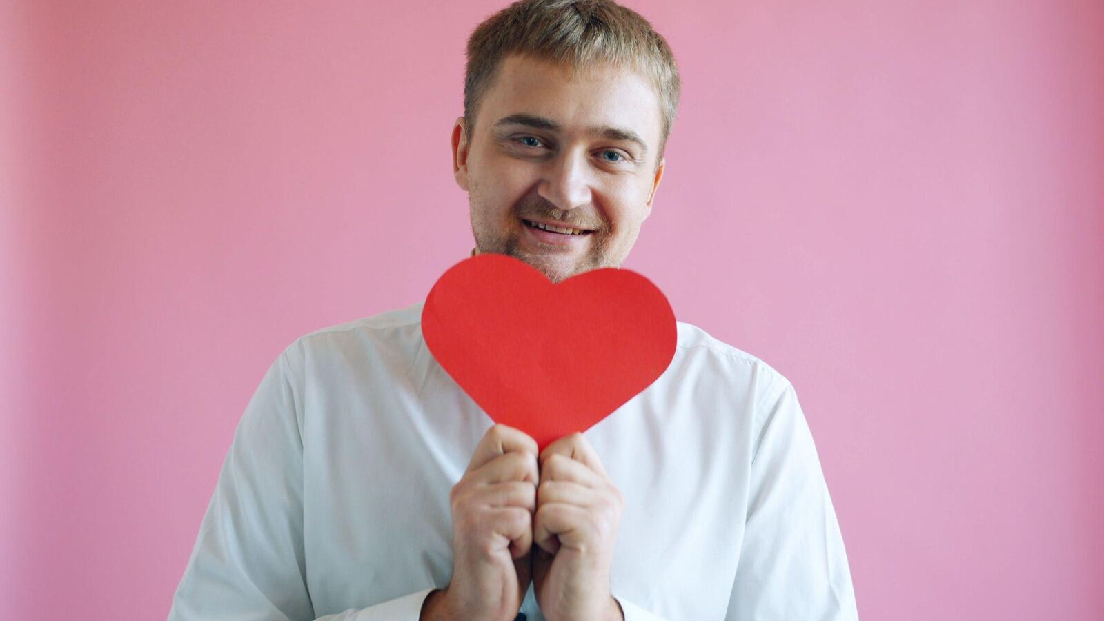 Ein Mann im weißen Hemd hält vor rosa Hintergrund ein rotes Papierherz vor sein Gesicht