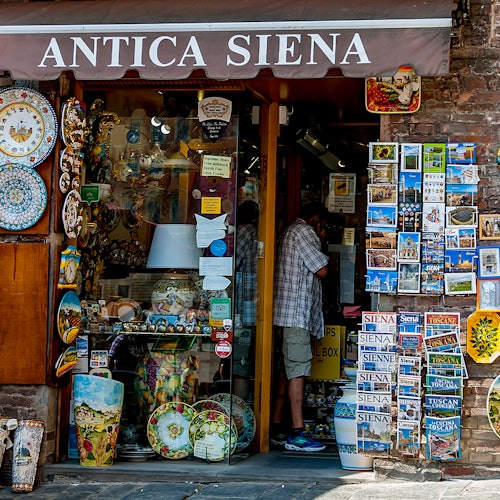 一家名为“Antica Siena”的商店，陈列着陶瓷盘、灯具和明信片。一位顾客站在店内浏览商品。