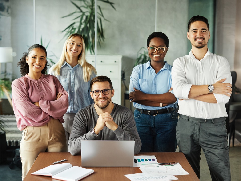 Grupo diverso de pessoas em pé, sorrindo e posando para a foto em frente a uma grande janela de escritório. A imagem transmite um ambiente acolhedor e colaborativo, refletindo a alegria e a união do grupo.