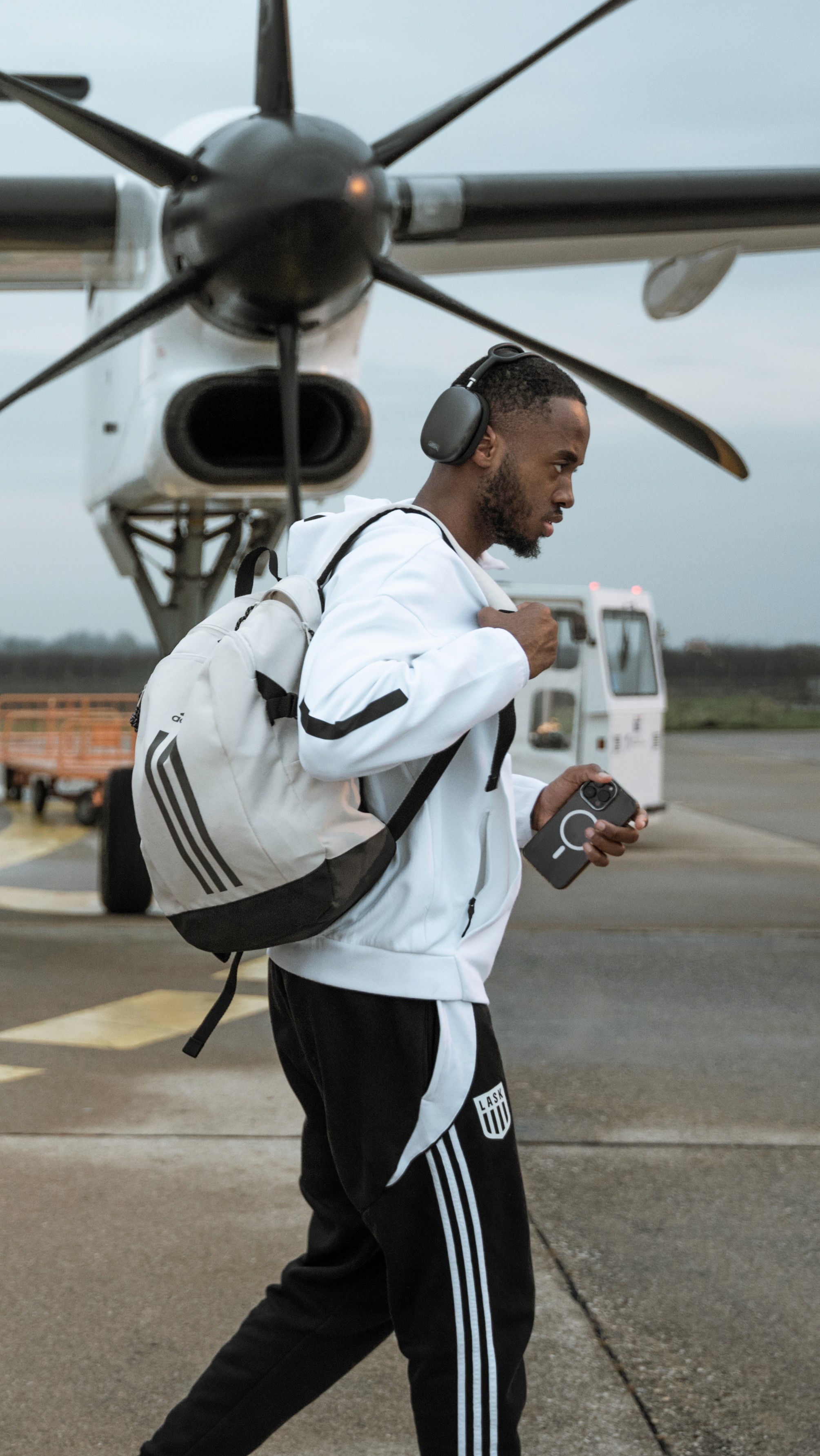 Ein Mann mit Kopfhörern und Rucksack, der an einem Flugzeug vorbeigeht und sein Handy hält.
