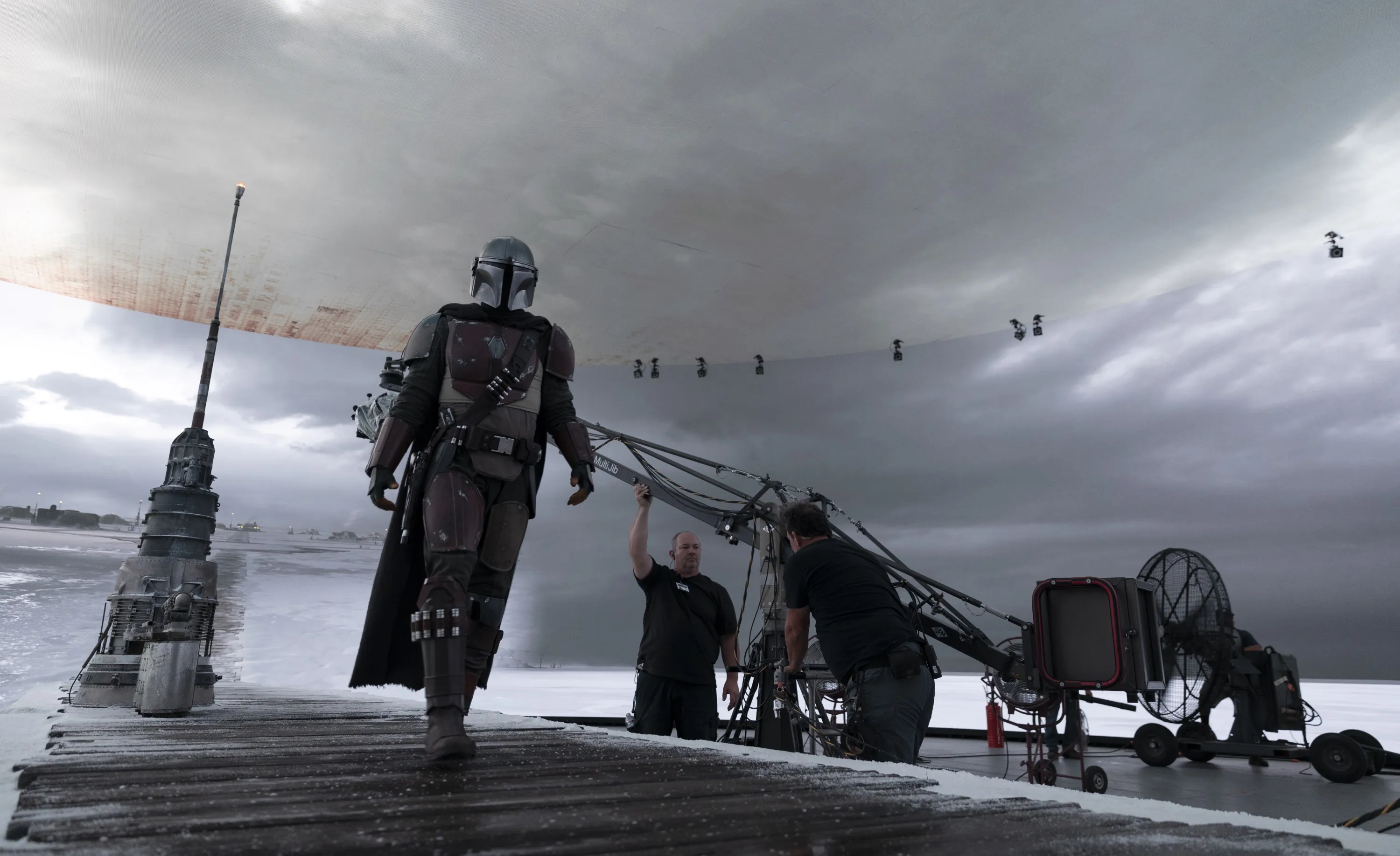 A person walks along a shore under dramatic, cloudy skies, with silhouetted figures and boats in the background.