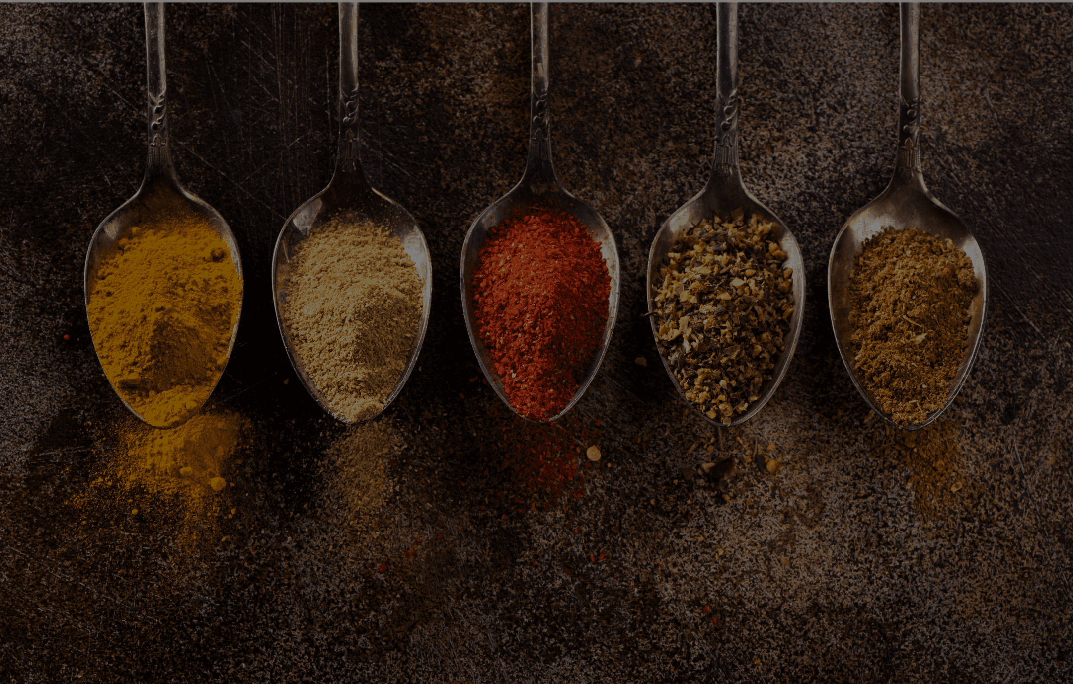 Assorted Indian spices on silver spoons against a rustic dark background, featuring turmeric, cumin, chili powder, mixed herbs, and garam masala. Perfect for Indian cuisine and spice blends.