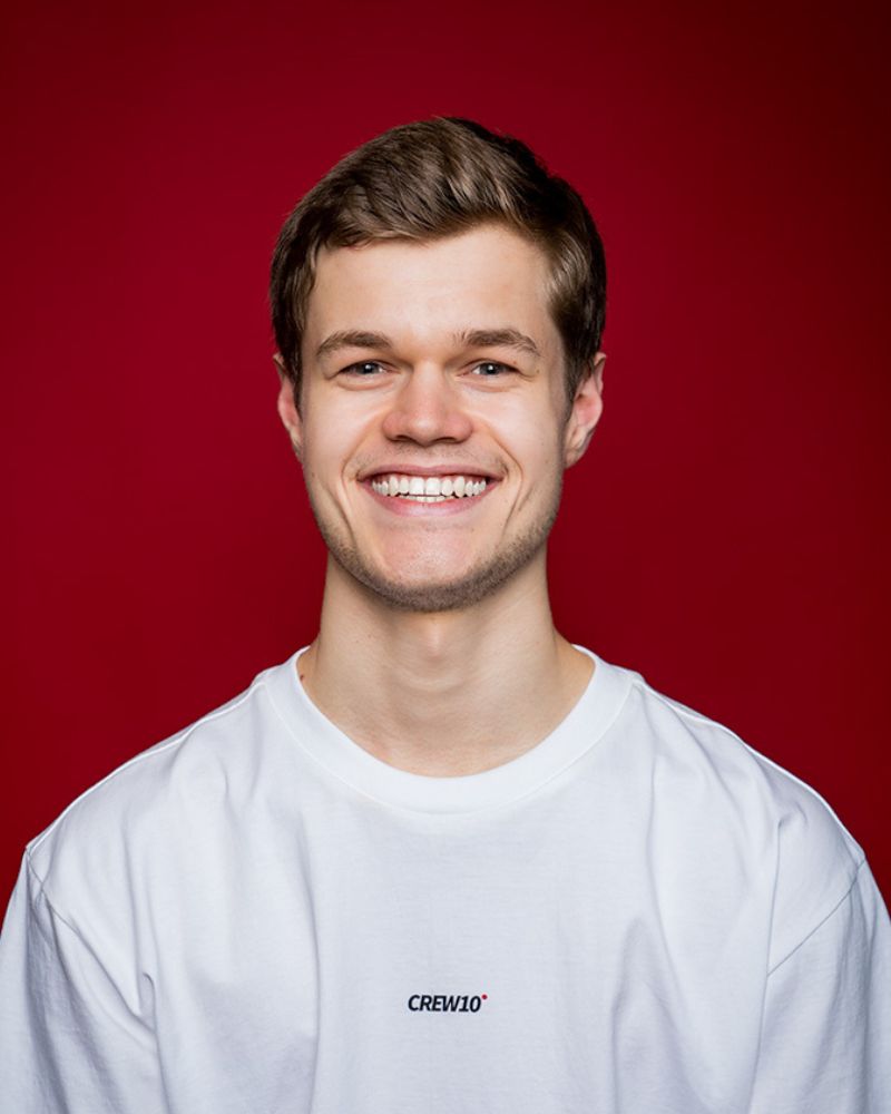 A smiling person with curly hair, wearing a leather jacket, stands in a cozy indoor setting.