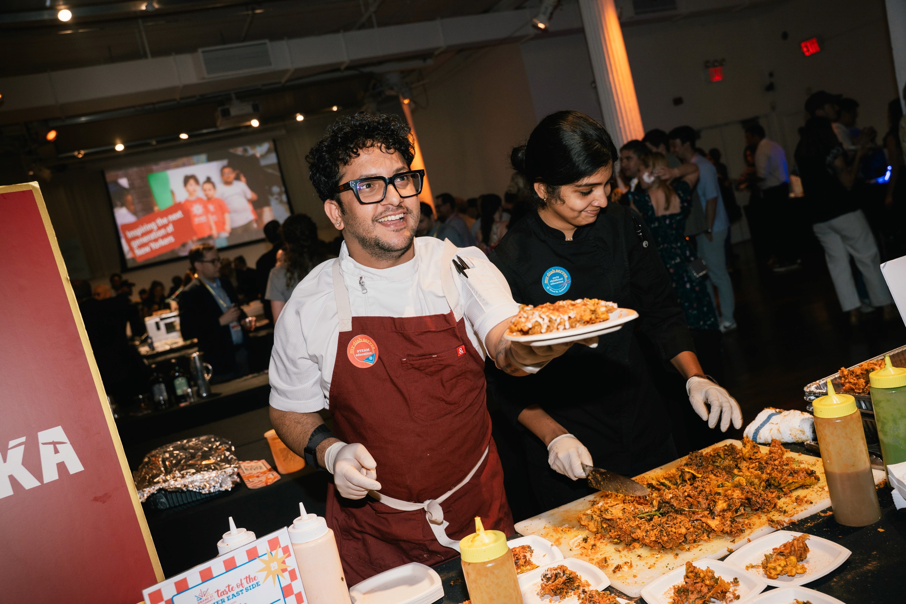A chef in a white shirt and maroon apron serving food inside of a bustling event space.