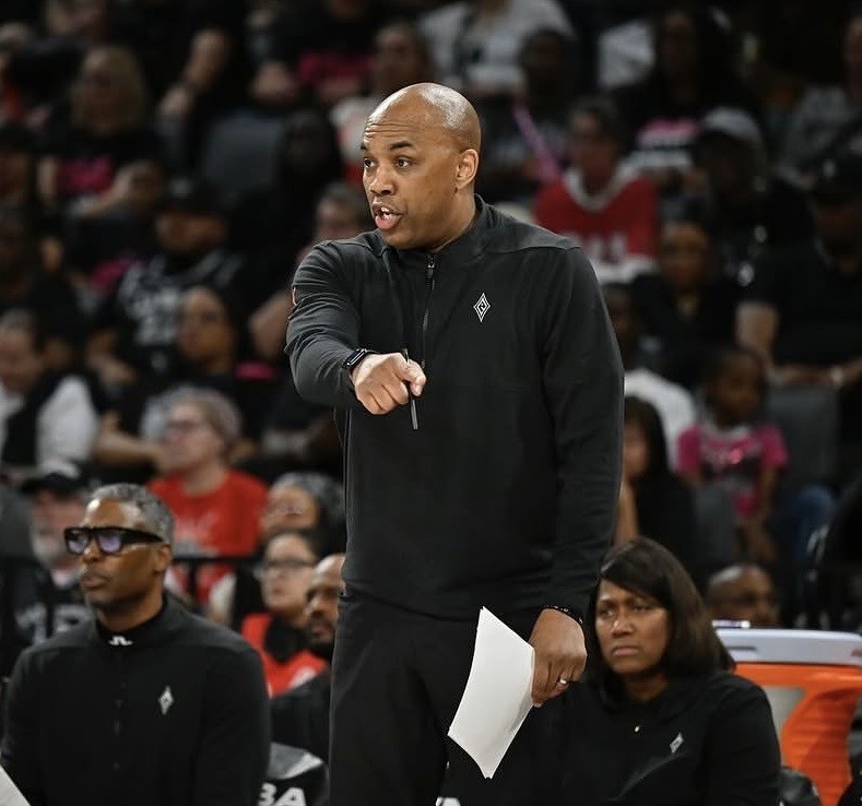 Coach Ty Ellis in a black tracksuit stands courtside, pointing and holding papers, while players and spectators watch intently in the background.