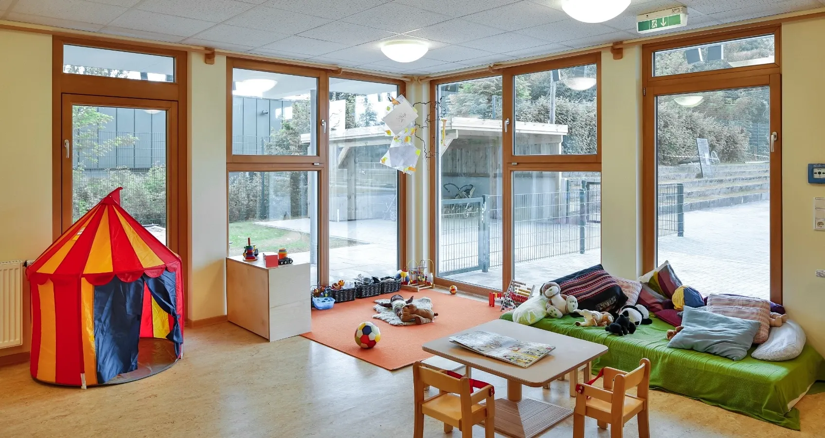 Spielzimmer mit Zirkuszelt, Kuscheltieren, Spielzeug und einem Sitzbereich mit vielen Kissen vor großen Fenstern.