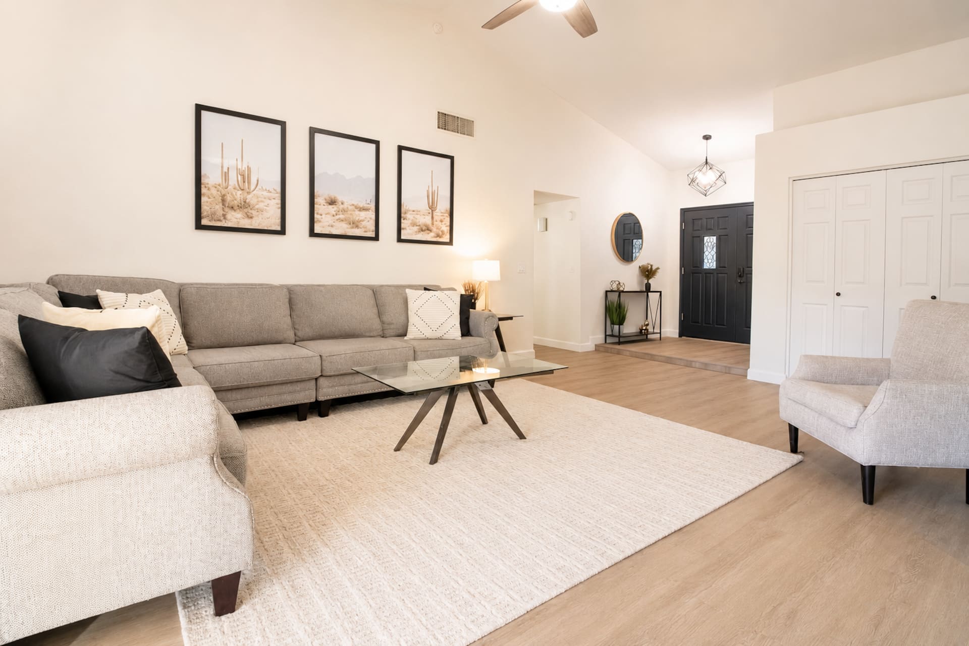 Open living room with L-shaped sectional sofa, glass coffee table, and hardwood floors. Cactus wall art and neutral decor. Connects to entryway with round mirror and console table.