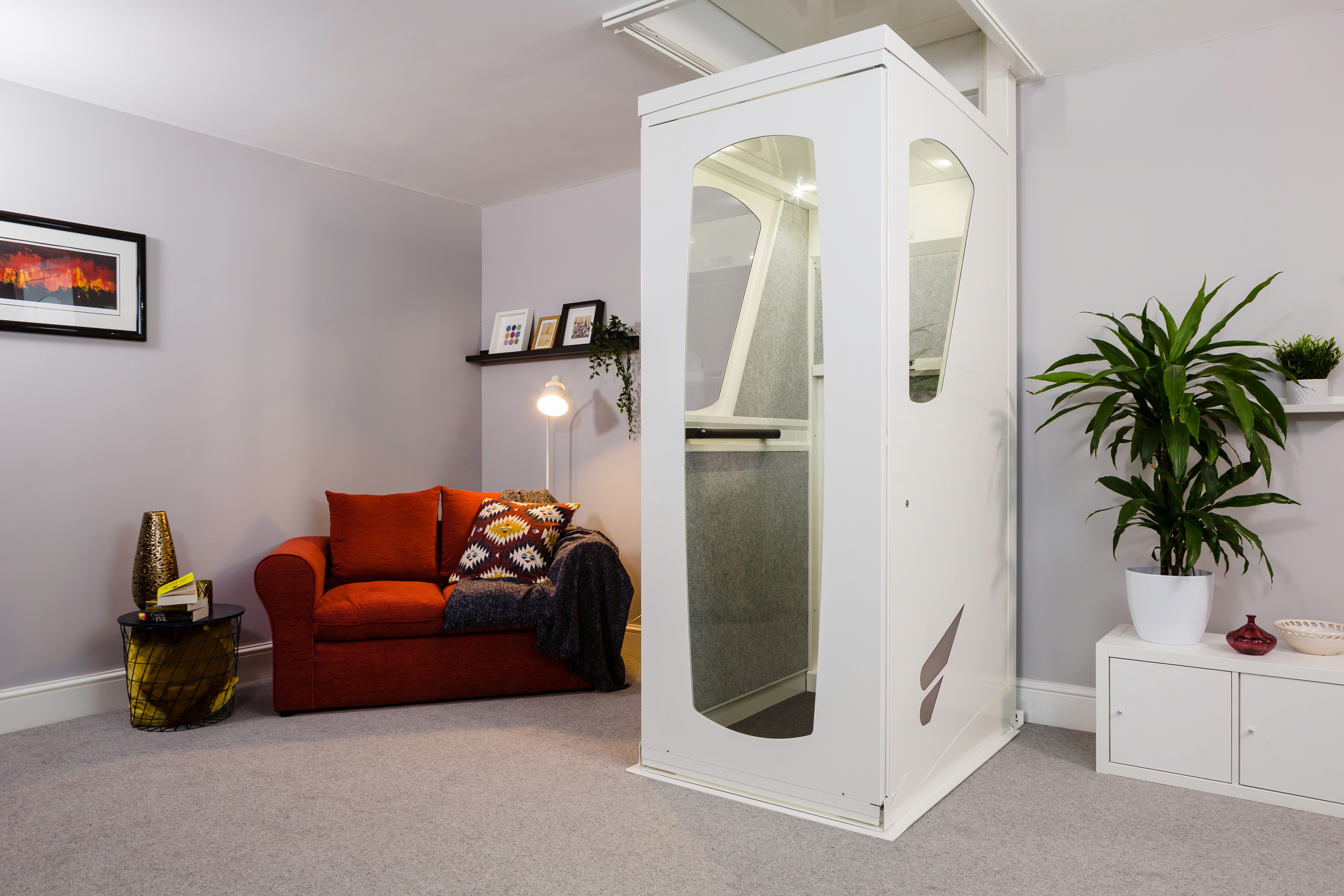 Elevate AVA home lift installed in a residential living room — white enclosed through-floor lift with internal lighting and glazed door panels, door closed