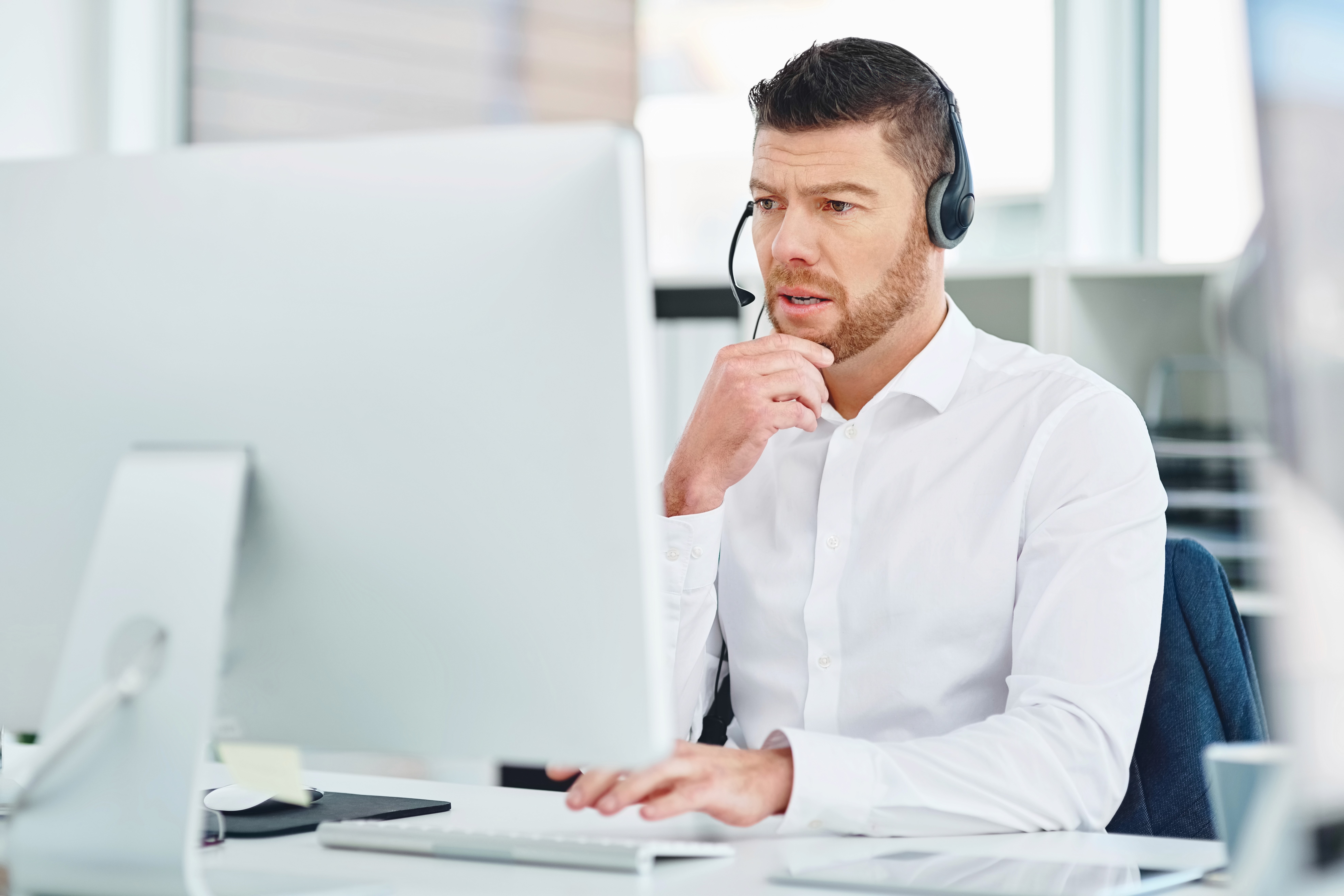 A man wearing a white shirt and headset works intently on a desktop computer in a bright, modern office setting.