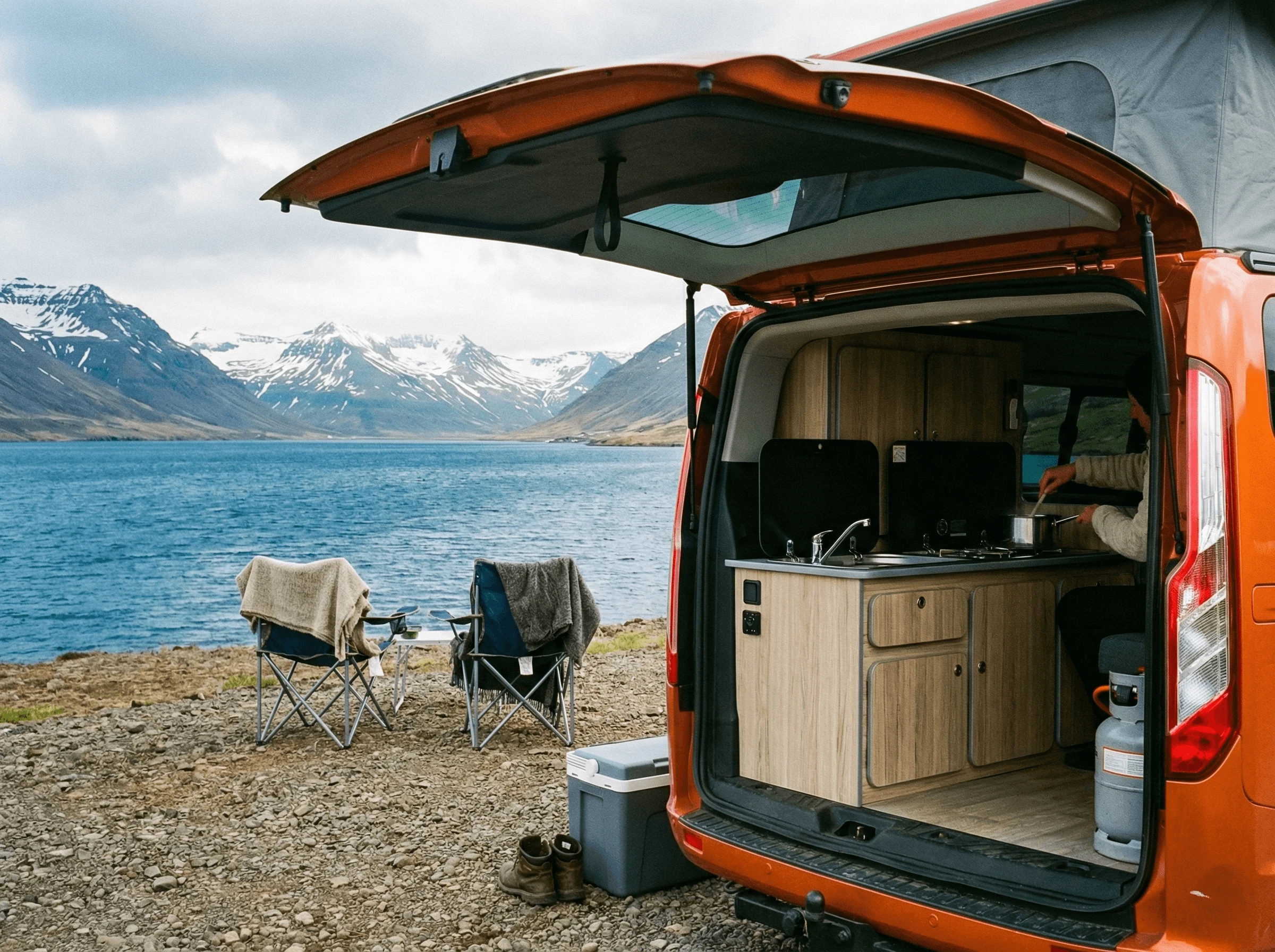 View from the open back of a camper van showing the wooden interior and camping chairs, overlooking a blue fjord and snowy mountains.