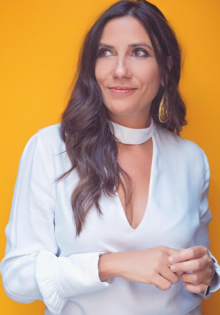 A young person with curly hair smiles at the camera while sitting in front of a laptop against a dark background.