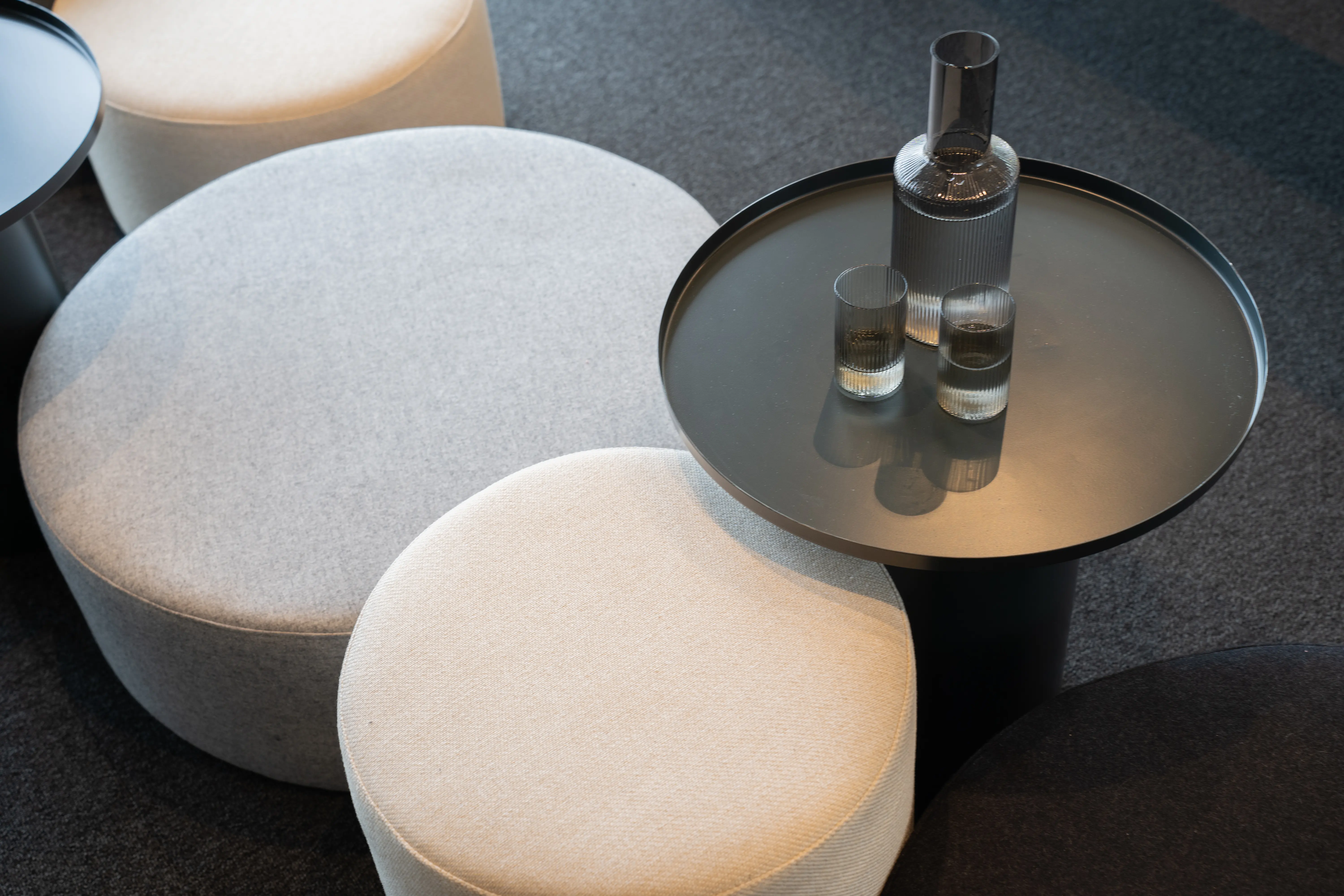 Top-down detail of round fabric poufs in cream and grey next to a black metal tray table with a water carafe and glasses at AI START-UP MUNICH.