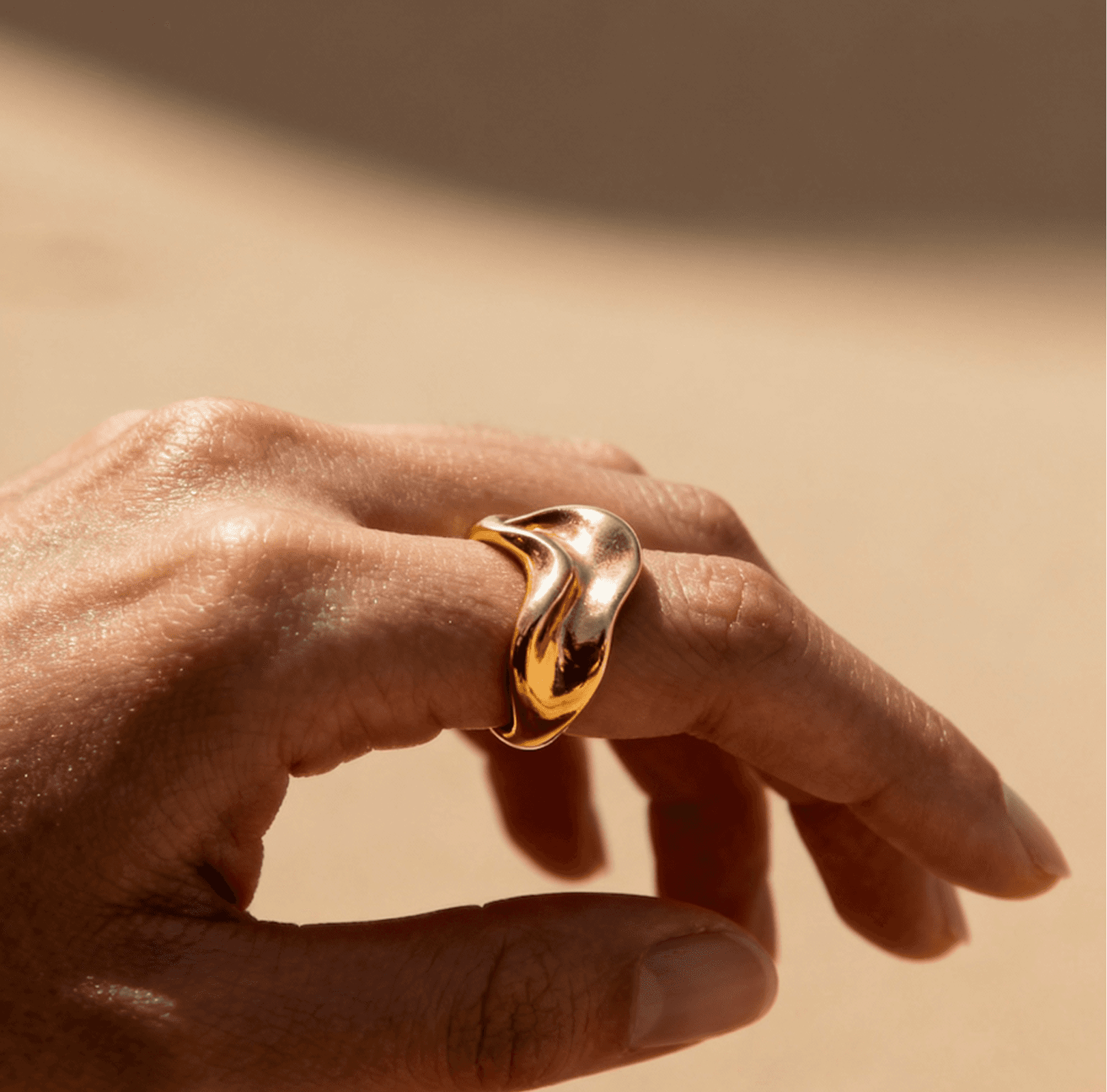 Close-up of a hand wearing a sculptural, wave-shaped gold ring in warm sunlight