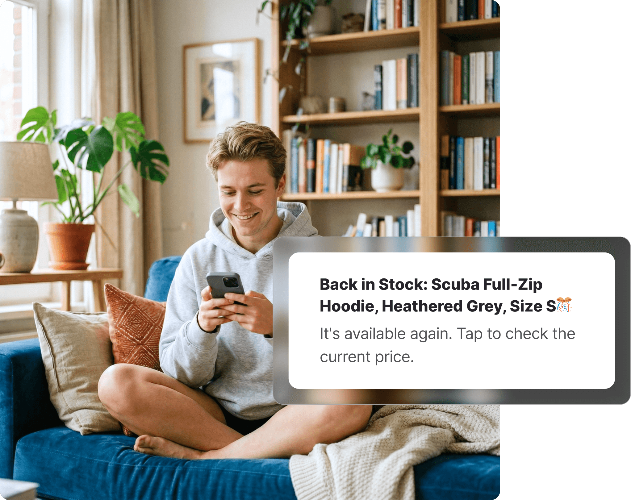 Young man in a grey hoodie sitting cross-legged on a blue couch checking his phone, with a lululemon stock alert notification visible, bookshelf and indoor plants in the background.