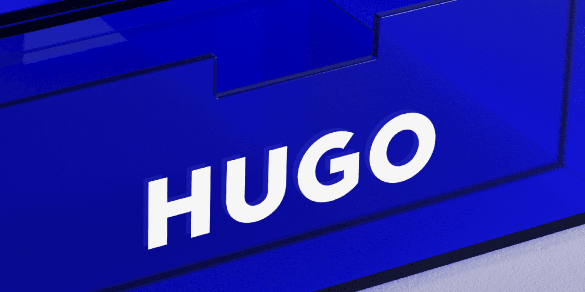 Markenwelt HUGO – Closeup der Acrylglas-Box in tiefem Hugo-Blau mit weisser „HUGO"-Wortmarke, Lichtreflexionen auf transluzenter Oberfläche.