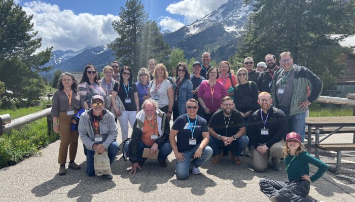 Group of about 30 people pose outside with snow-capped mountains and pine trees in the background. Most wear conference lanyards, and the weather is sunny. A child in front poses playfully on the ground.