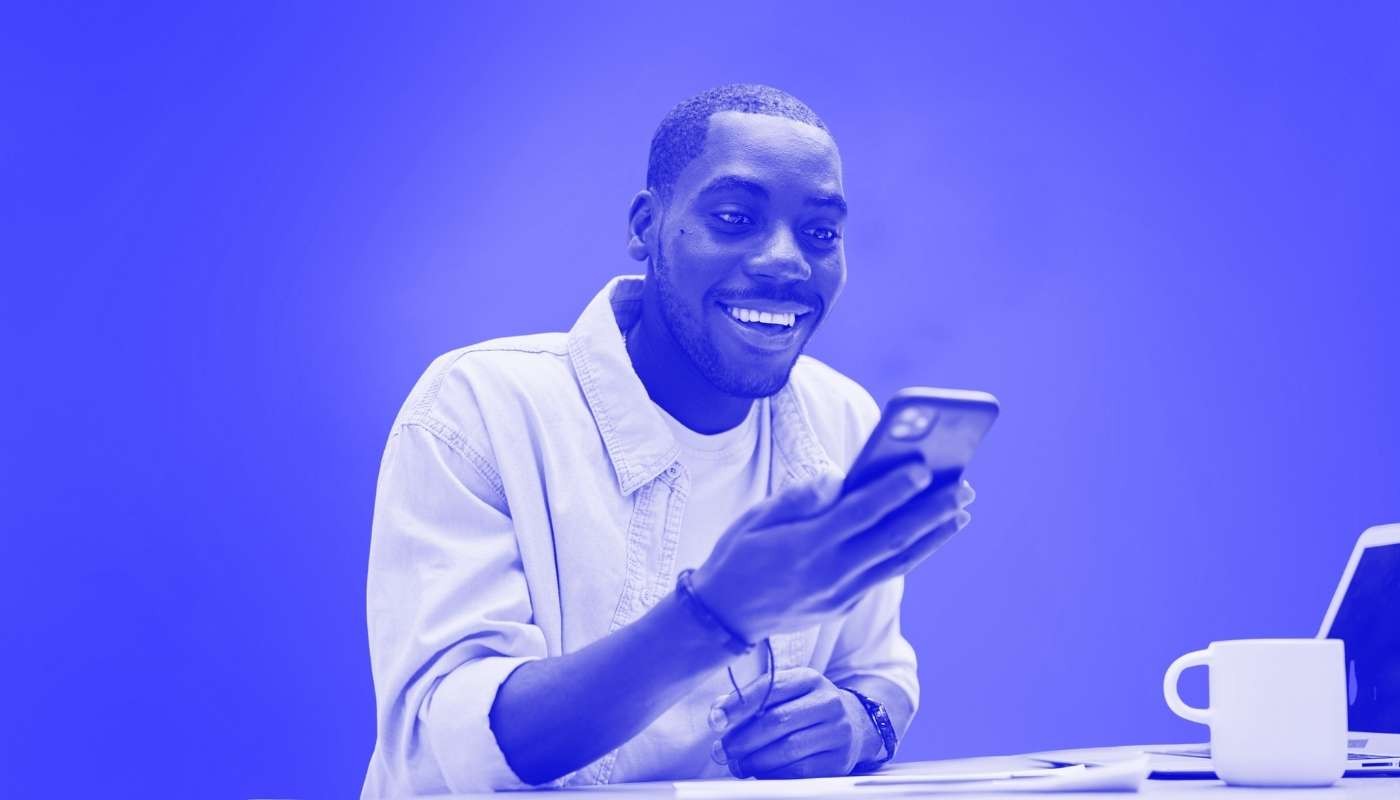 A man smiling while looking at his phone while sitting at a desk