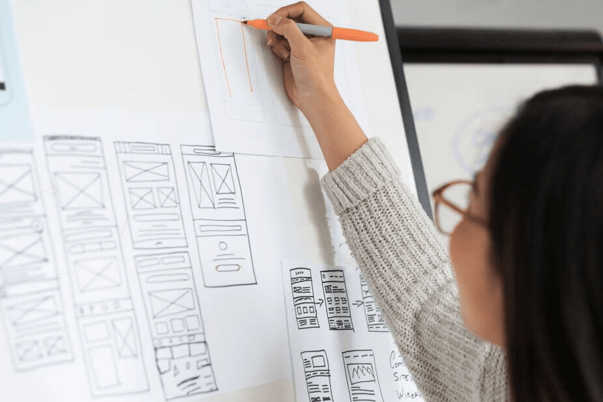 A woman with dark hair and glasses sketches wireframes out on a board