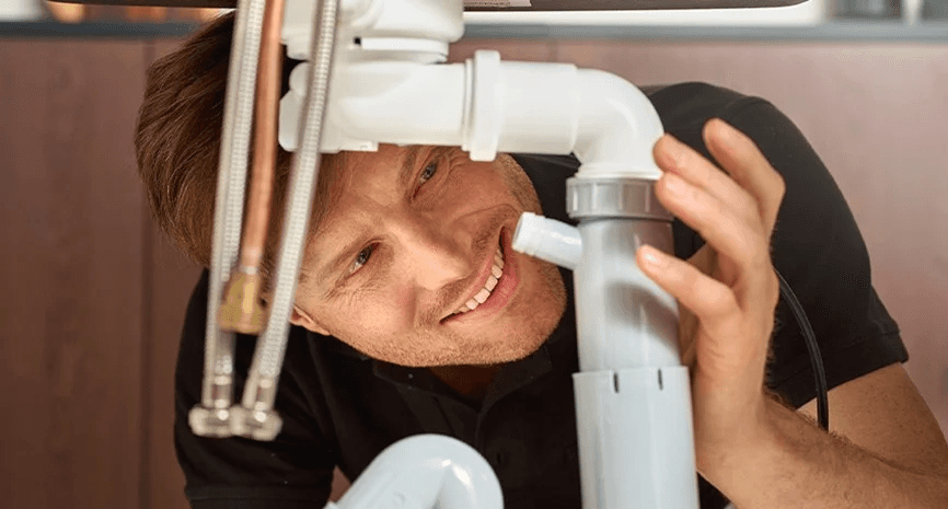 En rørlegger smiler mens han inspiserer rør under en vask