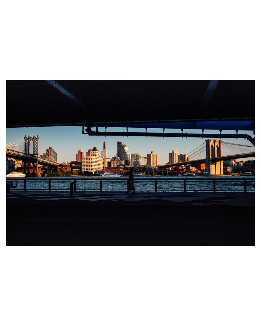 Photo of person walking under bridge with the east river, a view of Brooklyn including the Brooklyn Bridge in the background Photography