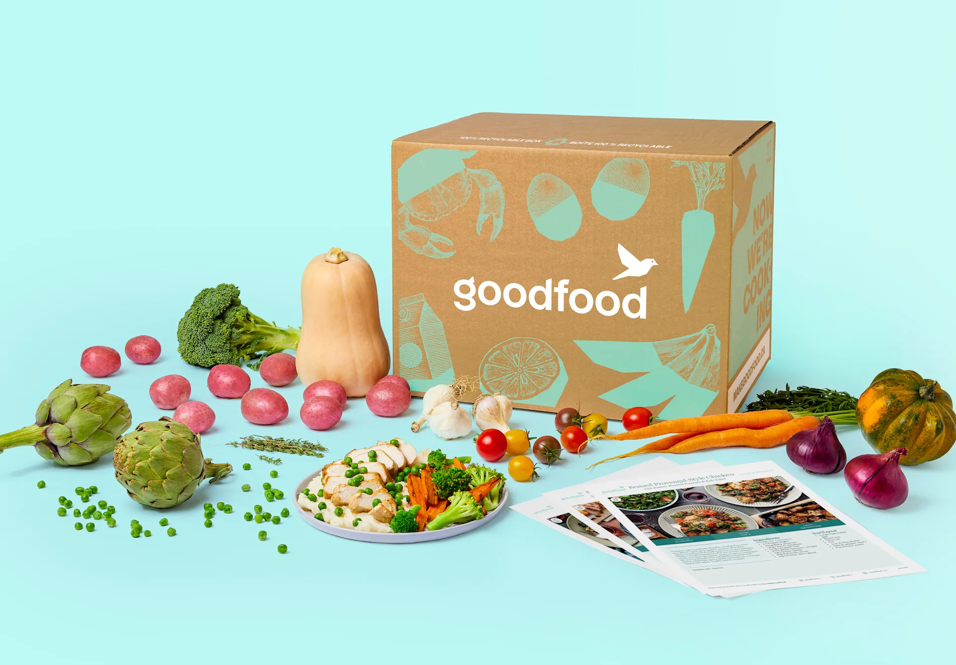 An open Goodfood meal kit delivery box on a blue background, surrounded by fresh raw produce like potatoes, broccoli, and carrots, alongside prepared meals and recipe cards.