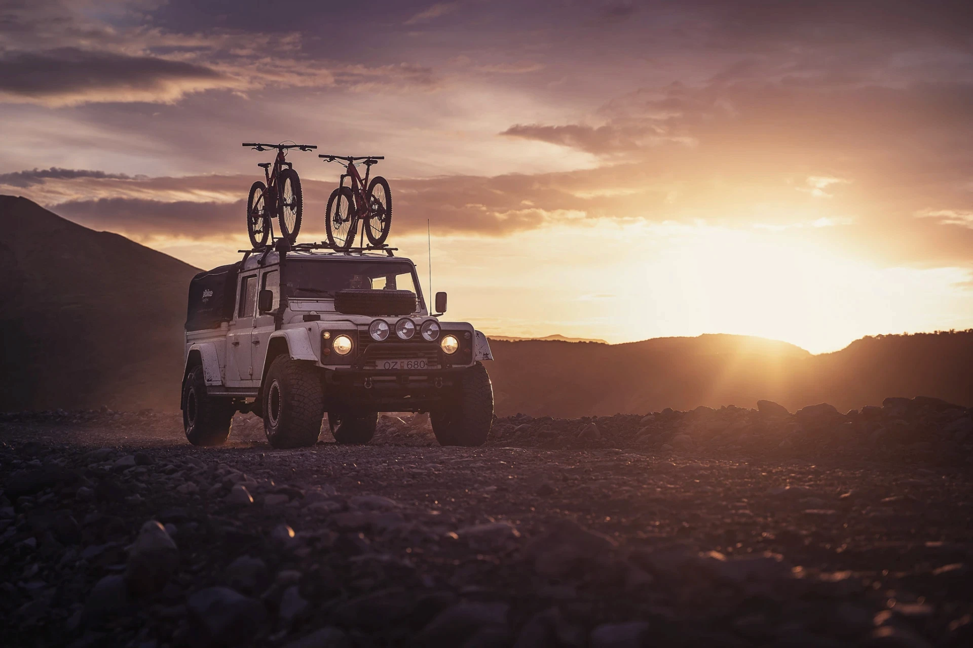 4x4 vehicle with two bikes on the roof parked on rocky ground at sunset.