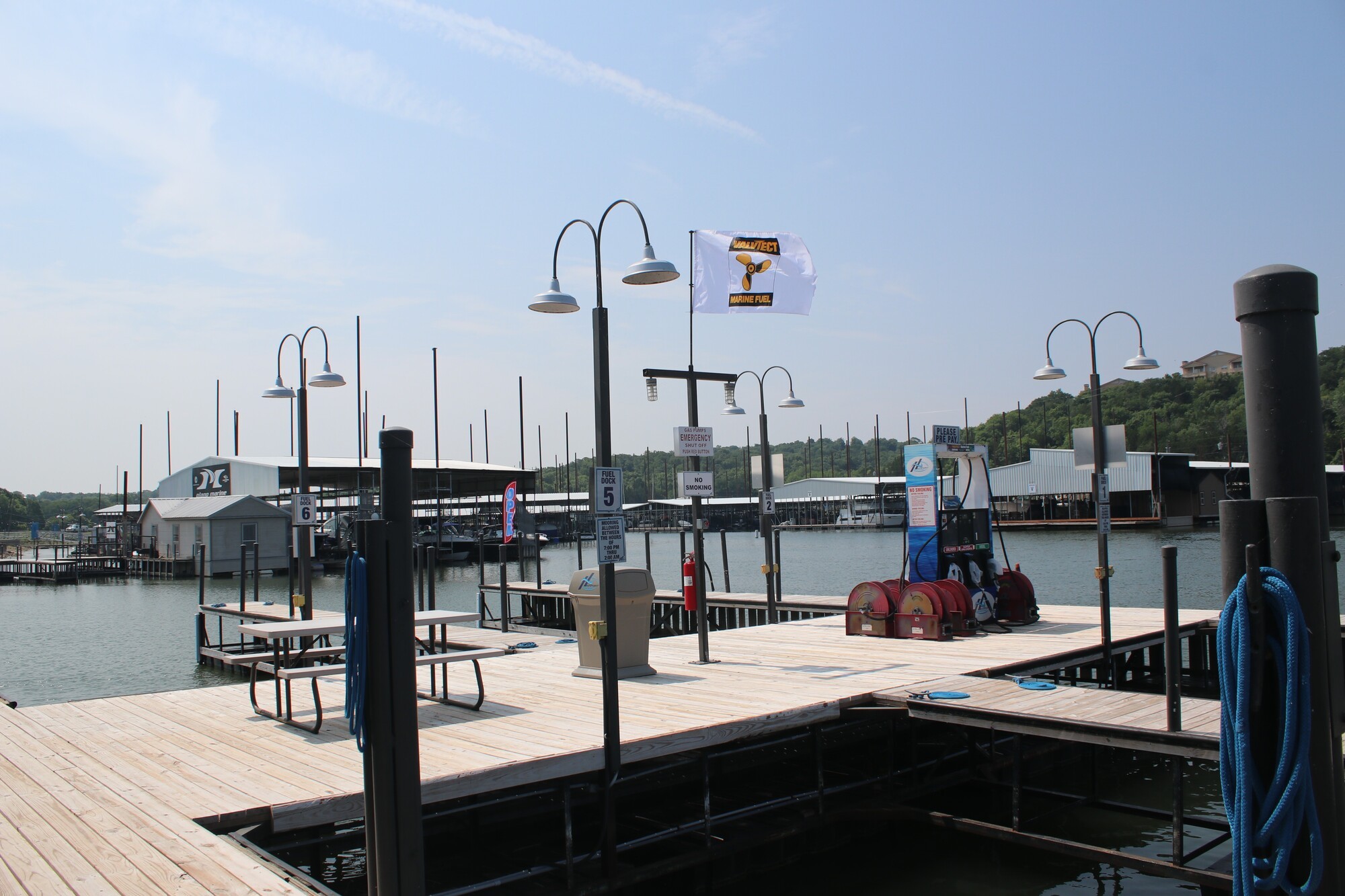 A tranquil marina scene showcases a dock equipped with mooring posts and boat fuel pumps, surrounded by covered boat slips and flanked by serene water under a clear blue sky.