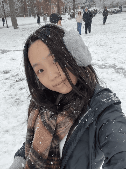 Una persona con una camiseta negra y pantalones de color claro está de pie en la cima rocosa de una montaña al atardecer, sosteniendo una cámara frente a su cara para tomar una foto. Un mar de nubes se extiende detrás de ellos, iluminado por la luz dorada del sol poniente.