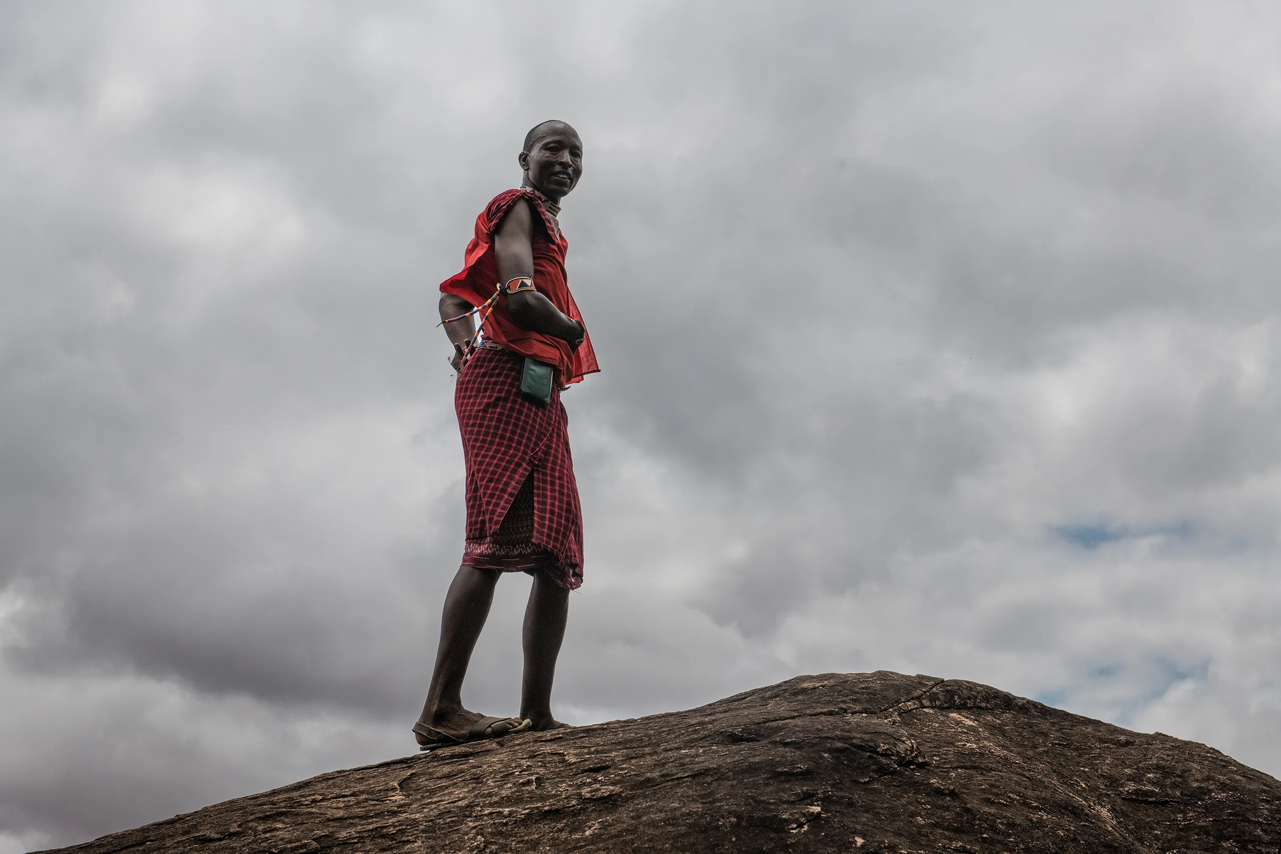 A Samburu warrior in traditional red attire standing on a rocky ledge overlooking the vast plains.