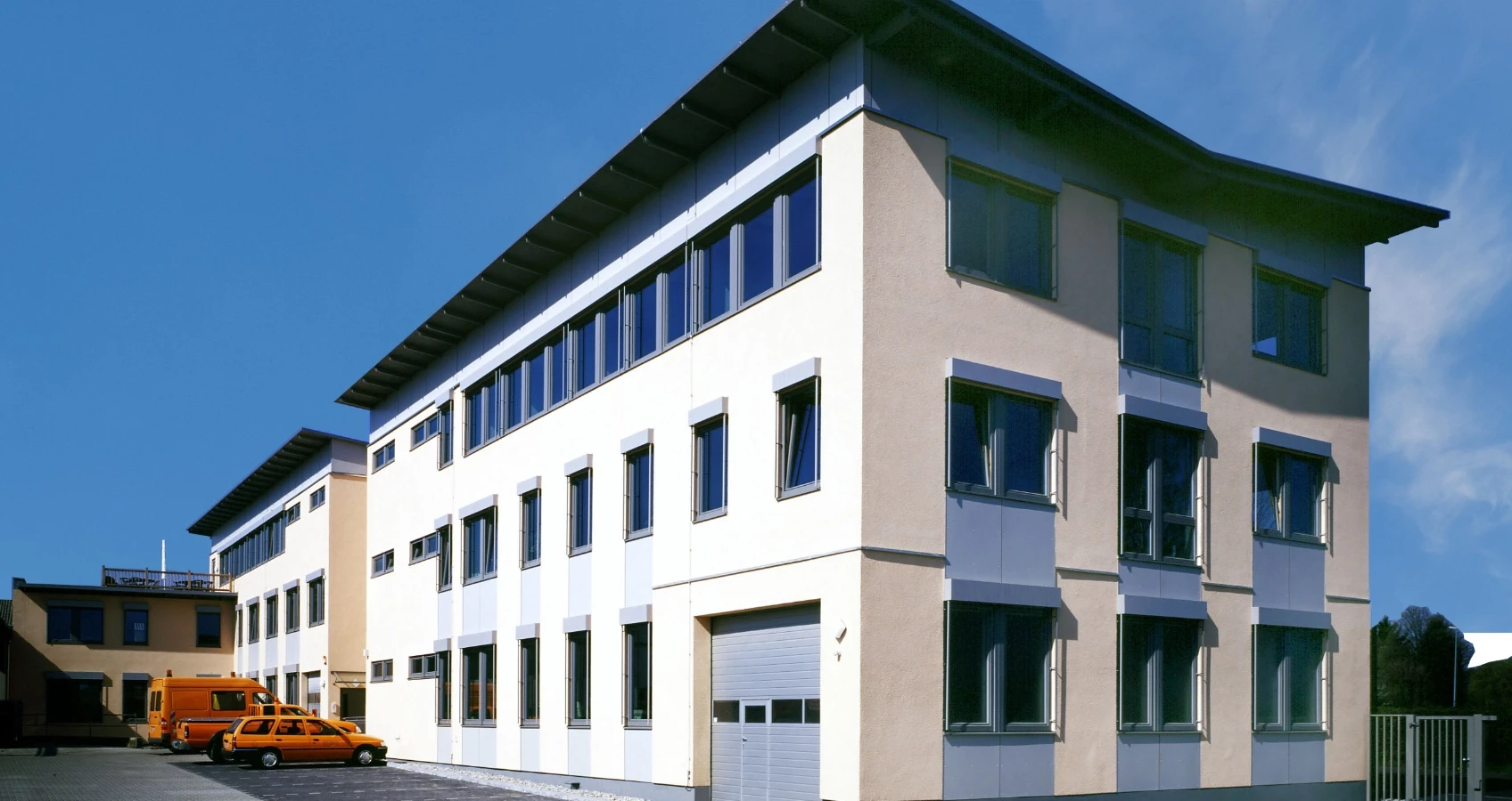 Moderne Bürogebäude mit vielen Fenstern und einer grauen Rolltor-Garage, im Hintergrund ein gelbes Fahrzeug und eine orangefarbene Limousine.