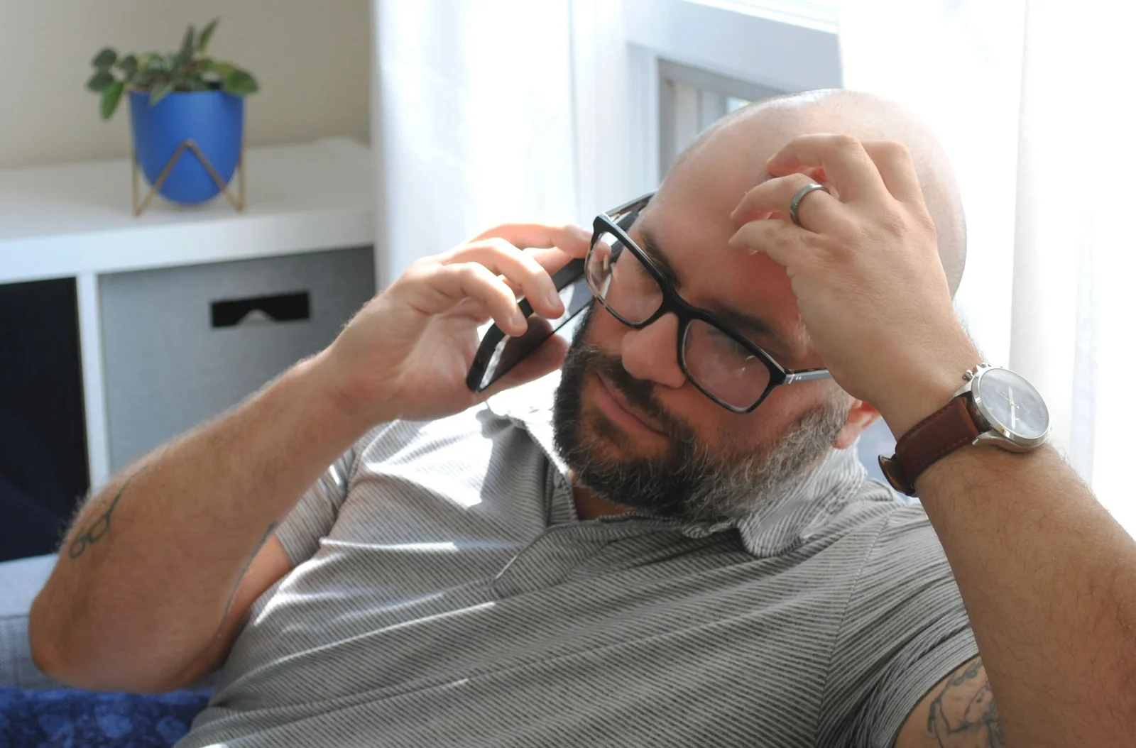 Man adjusting eyeglasses while on phone call in modern home office with potted plant visible.
