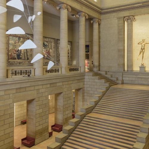 A grand staircase in a museum with columns, tapestries, a mobile sculpture, and a statue in the background.