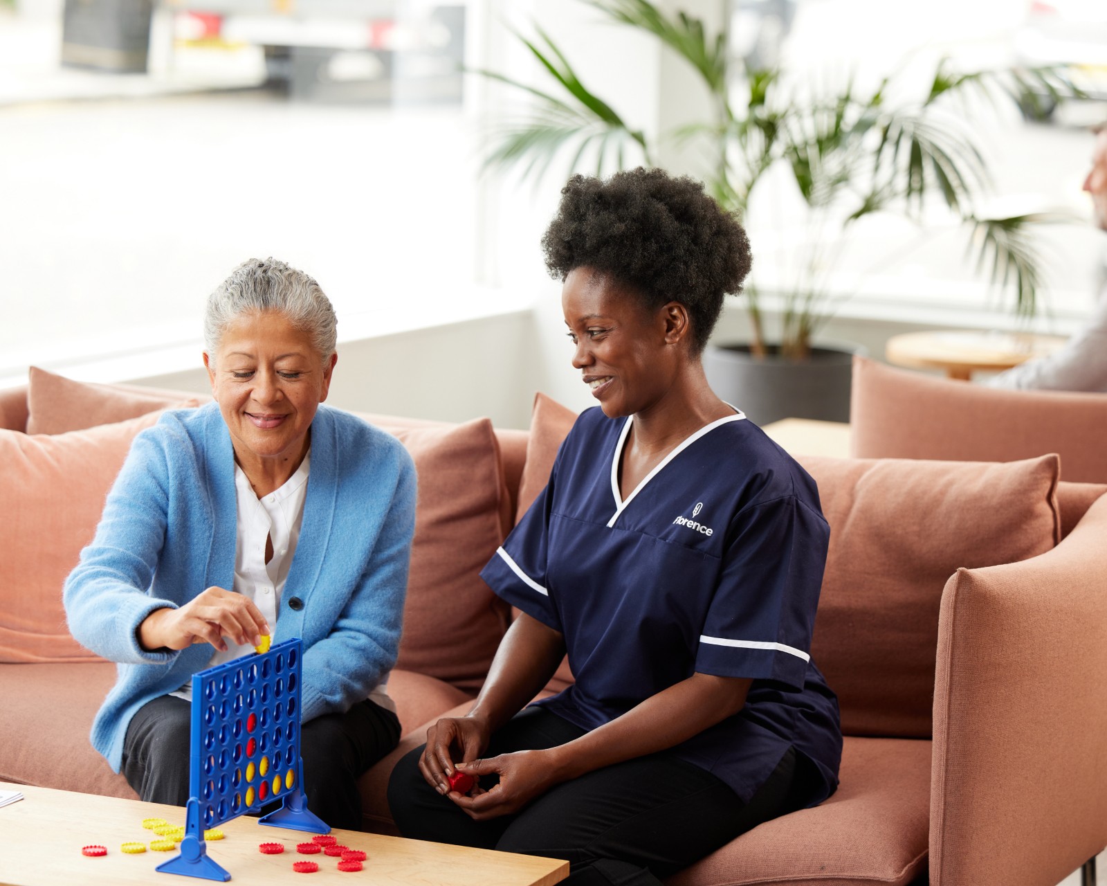 Older woman and Florence caregiver sitting on a sofa playing a board game together in a bright living room.