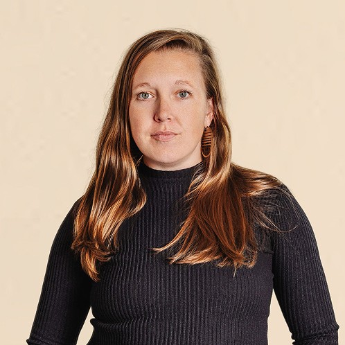 Woman with long hair wearing a black ribbed top, standing against a neutral background with a calm expression.