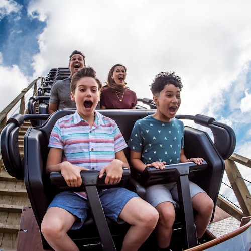 Two boys and two adults scream excitedly while riding a rollercoaster, with blue skies and clouds in the background.