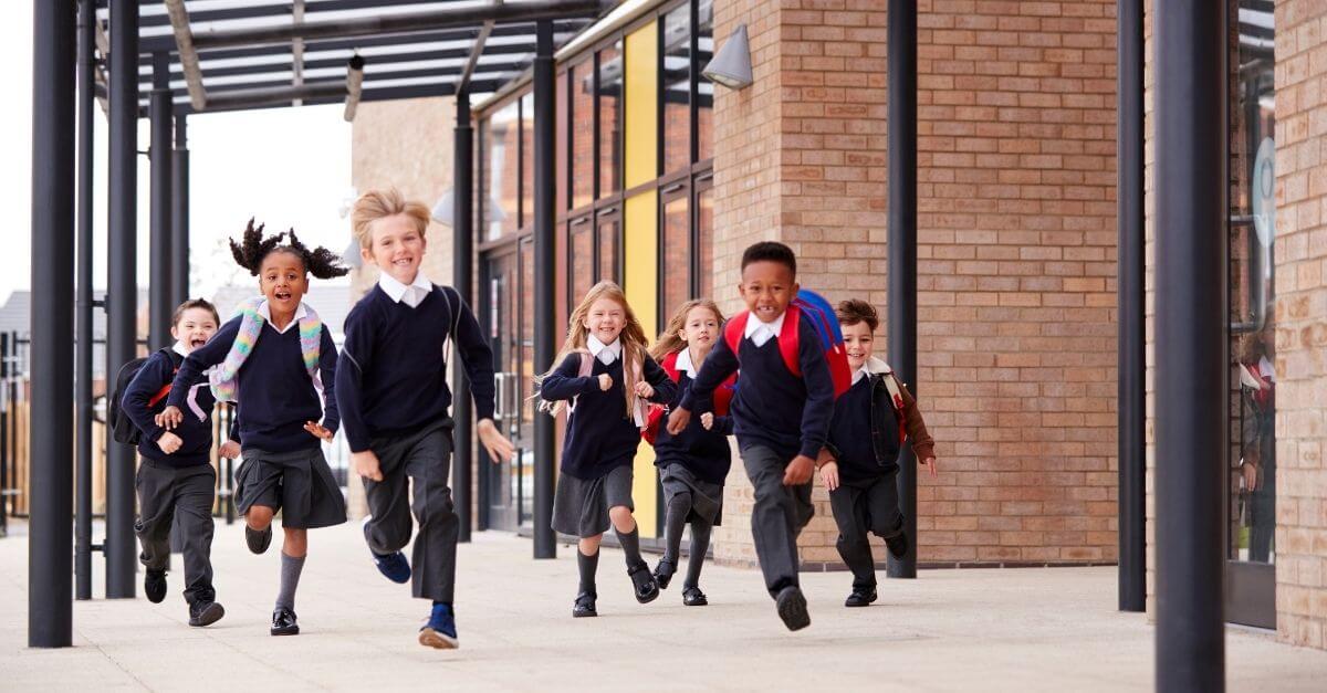 A diverse group of young schoolchildren in uniform running and smiling outside a modern school building.
