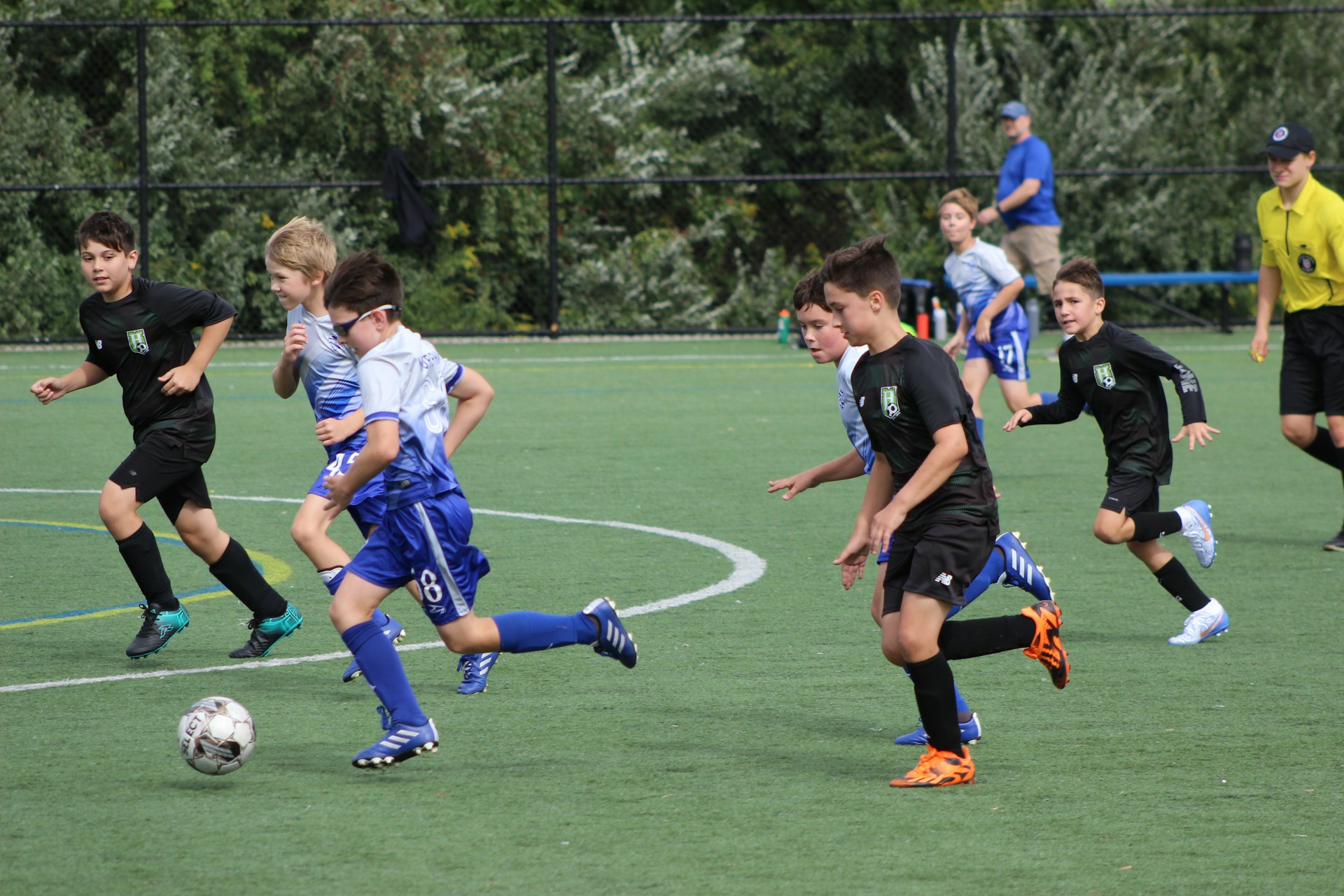 Young boys play a soccer match on artificial turf; a player in a blue kit dribbles the ball while being chased by opponents in black.