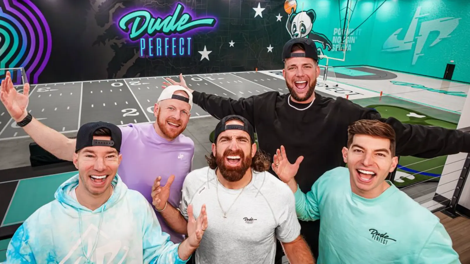 A group of five people smiling and posing together with arms raised in celebration against a colorful backdrop.