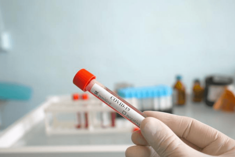 Gloved hand holding a test tube labeled ‘COVID-19’ in focus, with a blurred laboratory background featuring additional test tubes and bottles, representing medical testing.