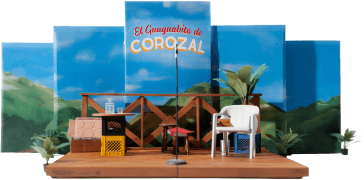 Maqueta del escenario, con un fondo pintado de montañas, ajones de leche plástico, una silla blanca plástica, cajas, plantas en tiesto, etc.