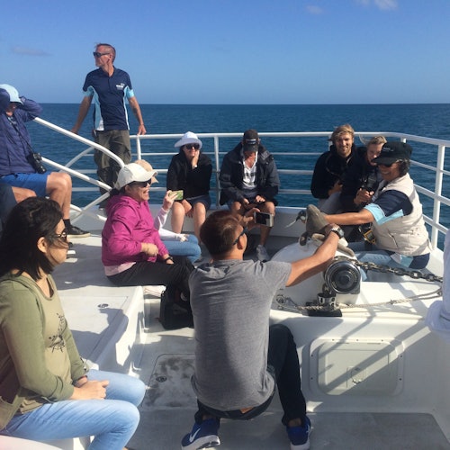 A group of people sits and stands on a boat's deck at sea, engaging in conversation under a clear blue sky.
